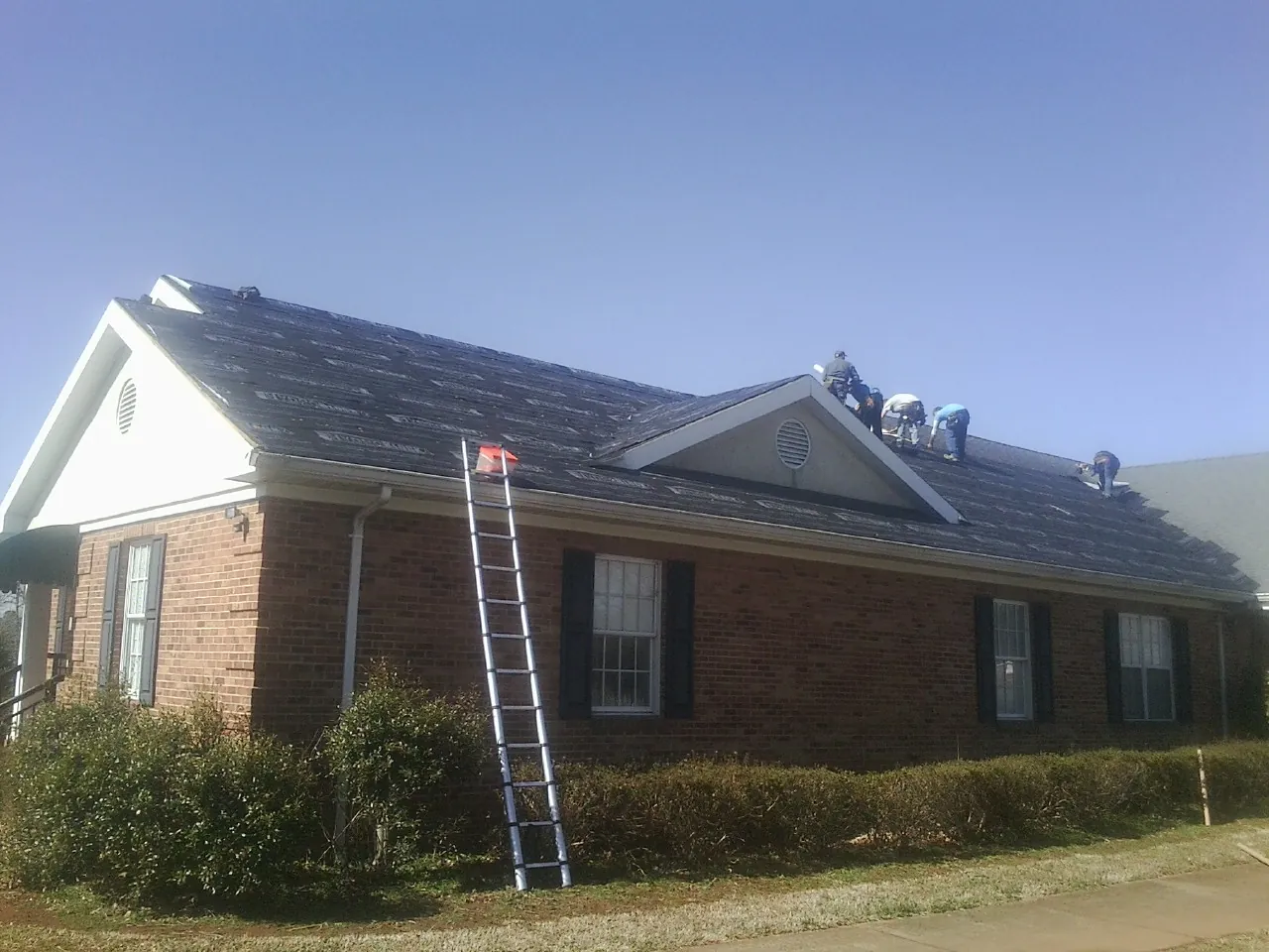 Crew tearing shingles off a brick ranch roof with a ladder set — Emergency Roof Repair work by our crew in Aredale