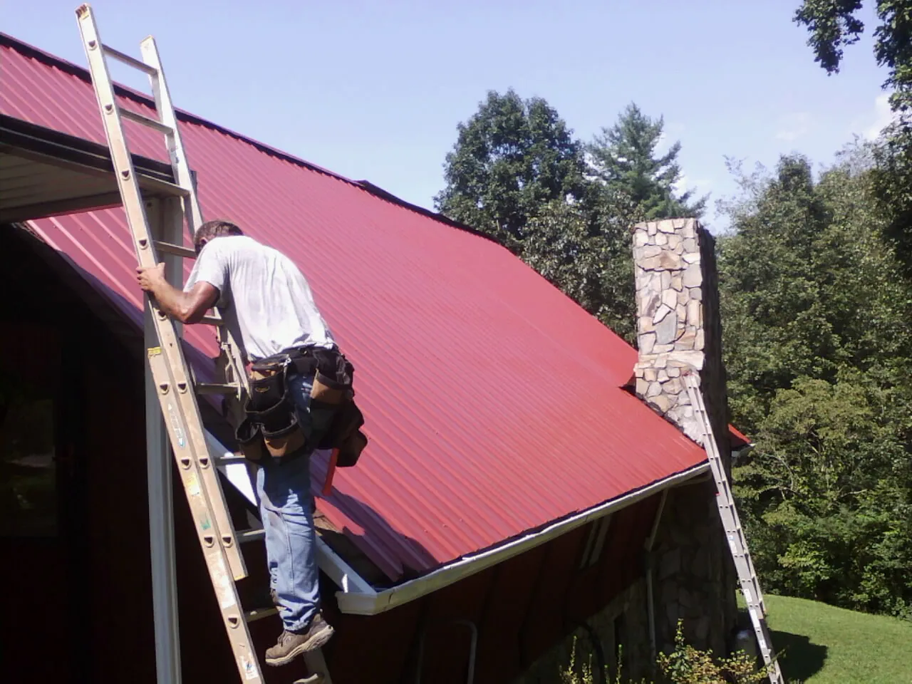 Roofer on a ladder beside a red metal roof and stone chimney — Soffit & Fascia Repair by Roofing Brothers in Aredale